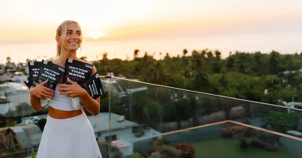Woman holding Better Than Water pouches on a rooftop with a sunset view